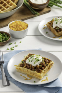 An image featuring a vertical view of golden cornbread waffles topped with a savory chili and a fresh sprinkle of shredded cheddar cheese and green onions. Real dairy provides a tangy and melty finish to a creative twist on a traditional meal and the high quality protein that adds a nutritional punch to every hearty bite.