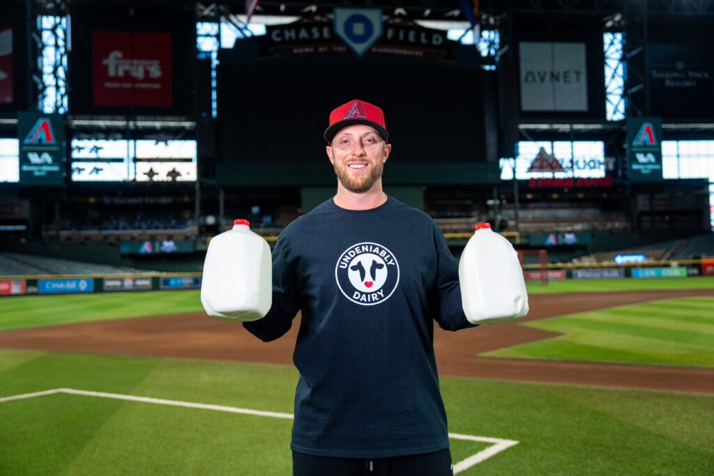 Dbacks pitcher Merrill Kelly on a baseball field holding two gallons of milk.